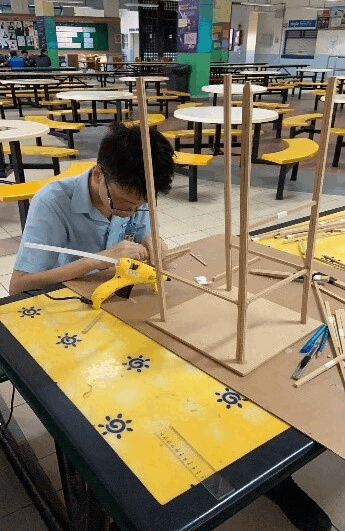 A student uses a hot glue gun to assemble a wooden stick structure on a yellow table in a school cafeteria.